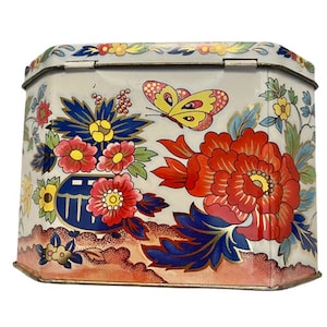 Può includere: Una scatola di latta vintage con sfondo bianco e un design floreale colorato. Il design presenta fiori rossi, gialli, blu e verdi, oltre a una farfalla gialla con segni neri.