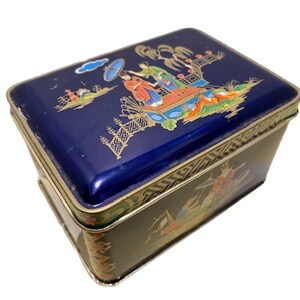 Può includere: Una scatola rettangolare in latta blu e oro con un design decorativo di ispirazione asiatica che presenta figure in abiti tradizionali e una pagoda. Il coperchio ha un bordo dorato e la scatola ha un bordo con motivi dorati e verdi.