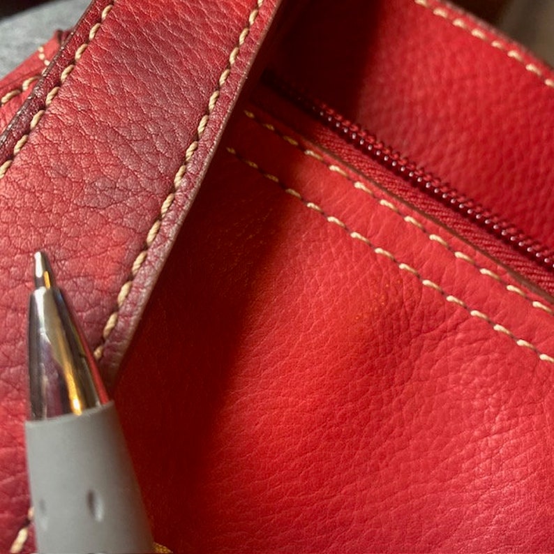 May include: Close-up of a red leather bag with white stitching and a zipper. The bag has a textured surface.