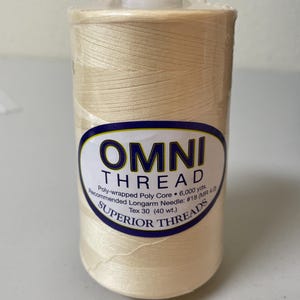 May include: A spool of cream-colored Omni Thread by Superior Threads, wrapped in clear plastic. The label reads: OMNI THREAD, Poly-wrapped Poly Core, 6,000 yds., Recommended Longarm Needle: #18 (MR 4.0), Tex 30 (40 wt.), SUPERIOR THREADS.