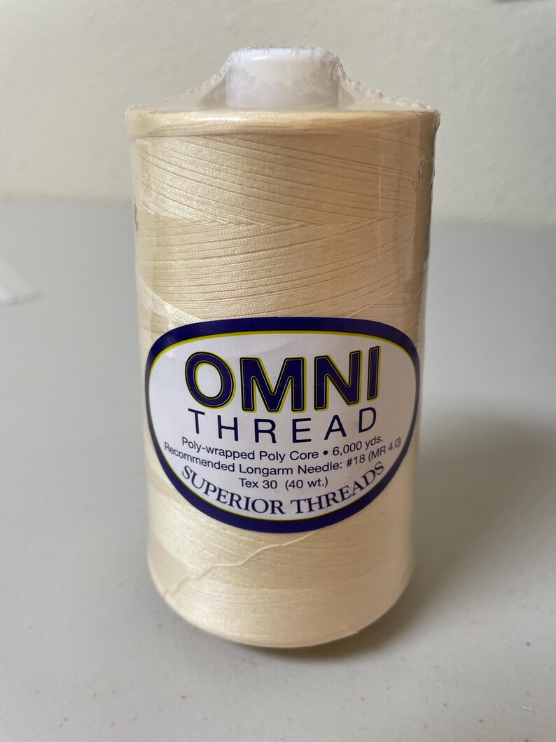 May include: A spool of cream-colored Omni Thread by Superior Threads, wrapped in clear plastic. The label reads: OMNI THREAD, Poly-wrapped Poly Core, 6,000 yds., Recommended Longarm Needle: #18 (MR 4.0), Tex 30 (40 wt.), SUPERIOR THREADS.