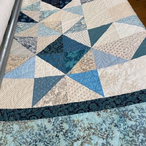 May include: A handmade quilt with a geometric pattern in shades of blue, cream, and tan. The quilt features various fabric textures and patterns, including floral and diamond shapes. The quilt is on a quilting frame.