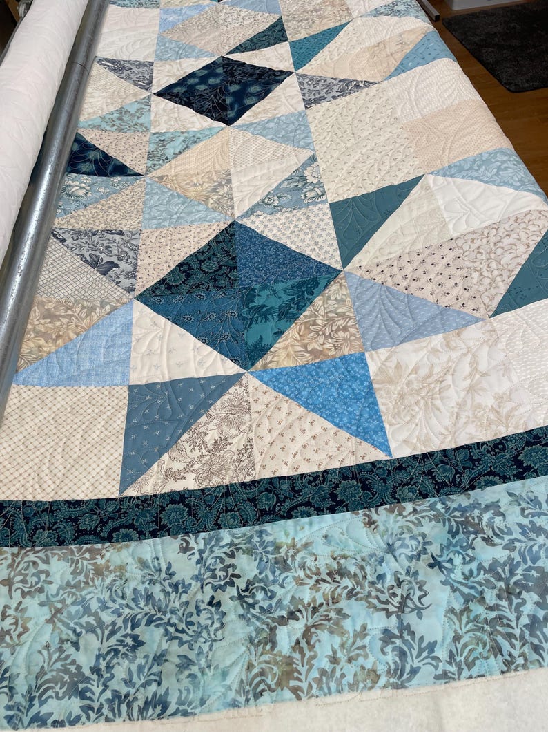 May include: A handmade quilt with a geometric pattern in shades of blue, cream, and tan. The quilt features various fabric textures and patterns, including floral and diamond shapes. The quilt is on a quilting frame.