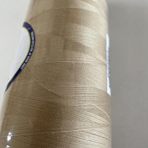 May include: A large spool of light tan sewing thread wound onto a cylindrical cone. A white label on the side features blue text that reads CONE MADE OF RECYCLED MATERIAL. The thread has a smooth, slightly reflective finish and is tightly wrapped.