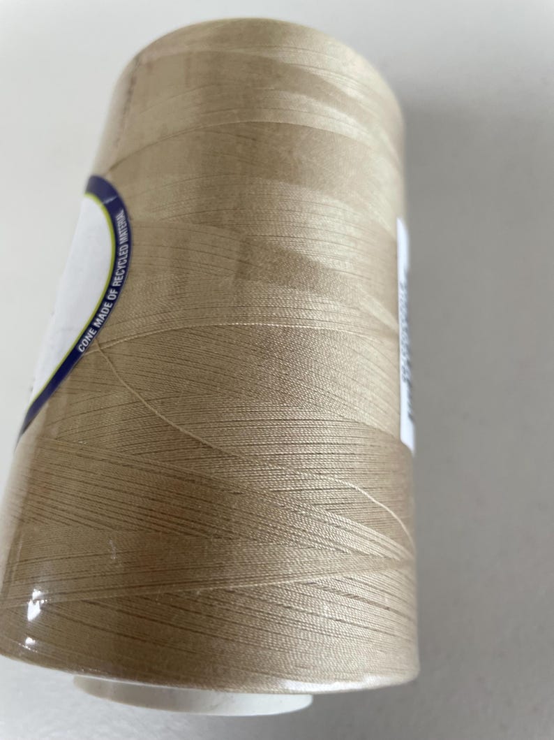May include: A large spool of light tan sewing thread wound onto a cylindrical cone. A white label on the side features blue text that reads CONE MADE OF RECYCLED MATERIAL. The thread has a smooth, slightly reflective finish and is tightly wrapped.