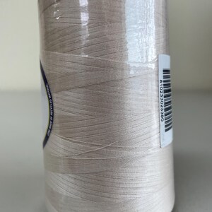 May include: A large spool of light beige thread wrapped in clear plastic film. A white label with a barcode and the number 810233023390 is attached to the side. A partial label with the text CONE MADE OF RECYCLED MATERIAL is visible.