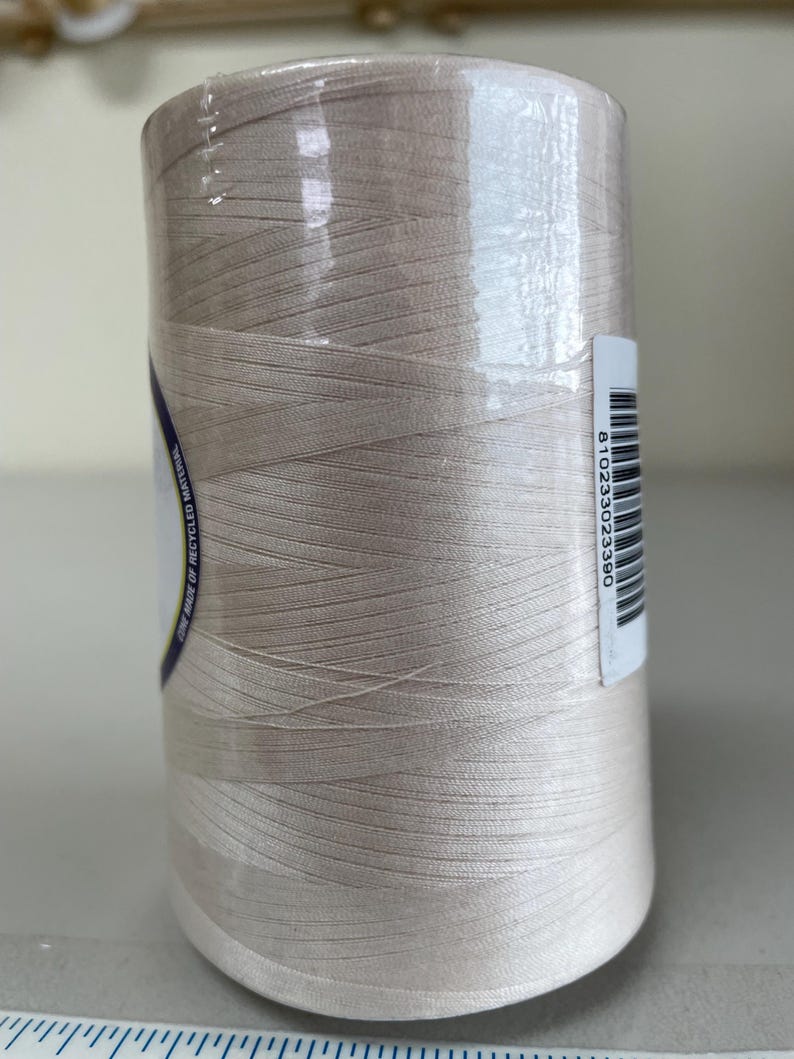 May include: A large spool of light beige thread wrapped in clear plastic film. A white label with a barcode and the number 810233023390 is attached to the side. A partial label with the text CONE MADE OF RECYCLED MATERIAL is visible.
