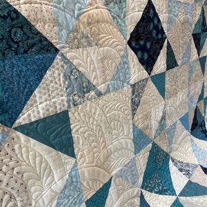 May include: A handmade quilt with a geometric design. The quilt features various shades of blue, cream, and white fabrics, arranged in a patchwork of triangles and diamonds. Intricate quilted patterns add texture and detail to the surface.
