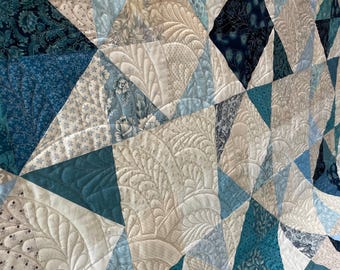 May include: A handmade quilt with a geometric design. The quilt features various shades of blue, cream, and white fabrics, arranged in a patchwork of triangles and diamonds. Intricate quilted patterns add texture and detail to the surface.
