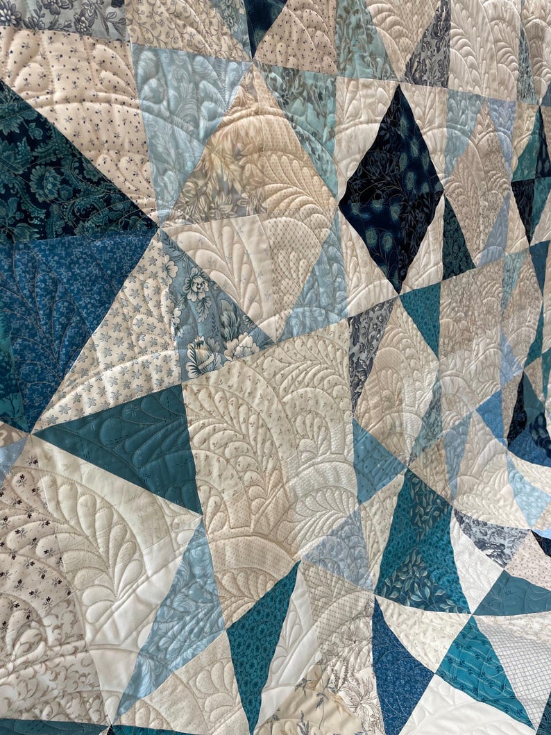 May include: A handmade quilt with a geometric design. The quilt features various shades of blue, cream, and white fabrics, arranged in a patchwork of triangles and diamonds. Intricate quilted patterns add texture and detail to the surface.