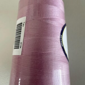May include: A large spool of light pink sewing thread wrapped in clear plastic packaging. A white label on the side features a barcode, the text "Tutu", the number "3134", and a circular label indicating "MADE IN VIETNAM" on the spool end.