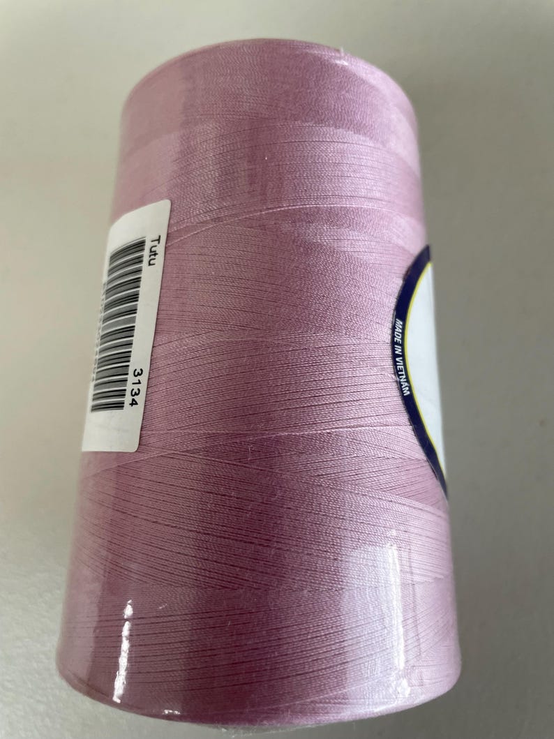 May include: A large spool of light pink sewing thread wrapped in clear plastic packaging. A white label on the side features a barcode, the text "Tutu", the number "3134", and a circular label indicating "MADE IN VIETNAM" on the spool end.