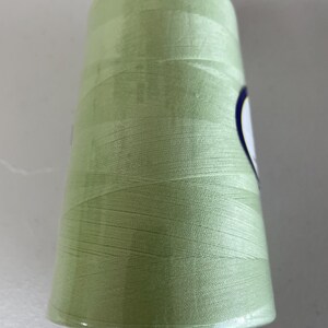 May include: A large spool of light sage green sewing thread. The thread is wound tightly around a cylindrical plastic core, creating a smooth, uniform surface texture. A small portion of a white label with a dark blue border is visible on the side.
