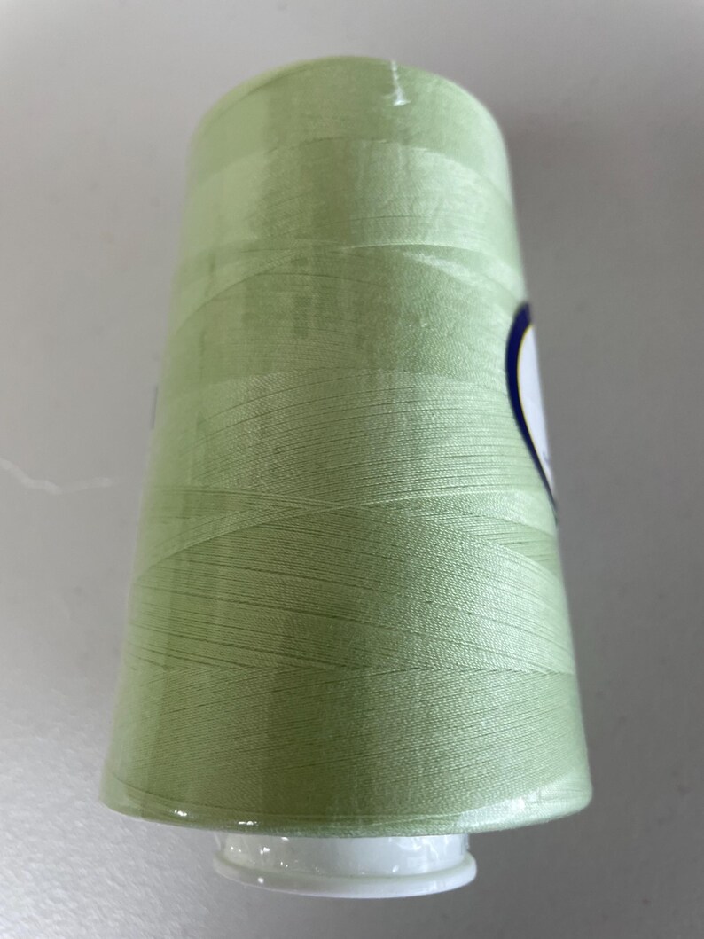 May include: A large spool of light sage green sewing thread. The thread is wound tightly around a cylindrical plastic core, creating a smooth, uniform surface texture. A small portion of a white label with a dark blue border is visible on the side.