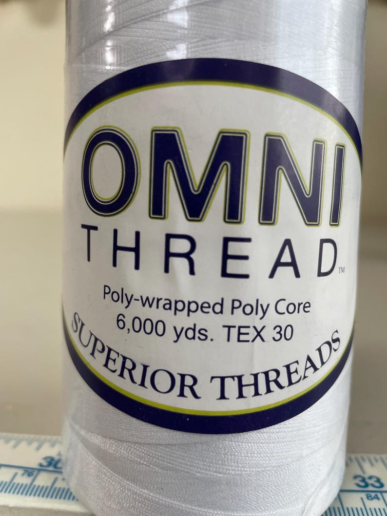 May include: A large spool of white Omni Thread with a label reading OMNI THREAD, Poly-wrapped Poly Core, 6,000 yds. TEX 30, and SUPERIOR THREADS. The spool is positioned against a measuring tape showing markings in inches and centimeters.