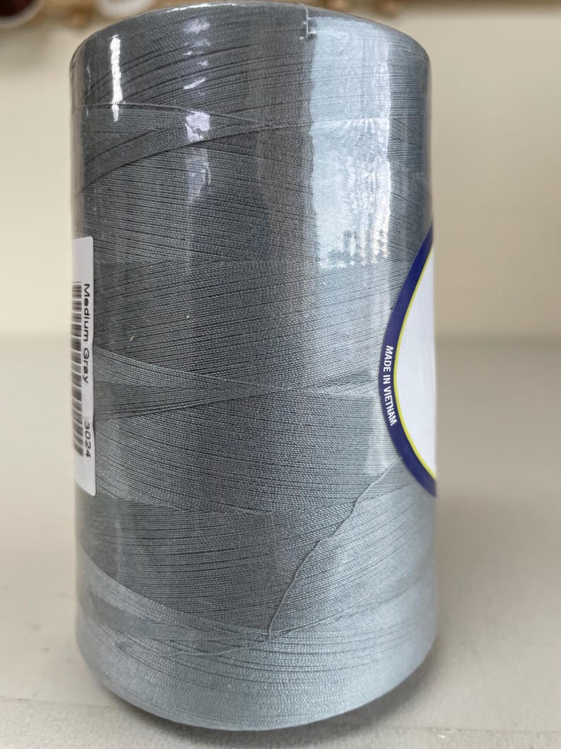 May include: A large cylindrical spool of medium gray sewing thread wrapped in clear plastic film. A white label on the side displays the text Medium Gray and 3024, while another label on the right side includes the text Made in Vietnam.