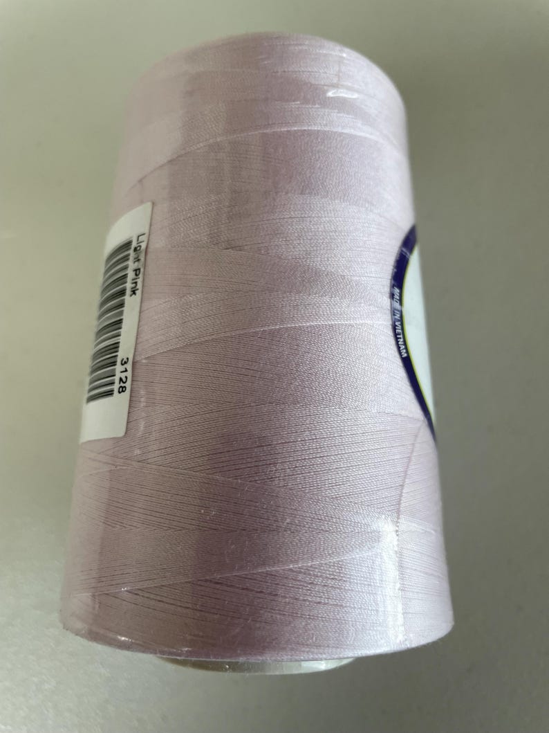May include: A large spool of light pink sewing thread wrapped in clear plastic film. A white rectangular label is attached to the side, featuring a barcode, the text Light Pink, and the number 3128. A partial label reads Made in Vietnam.