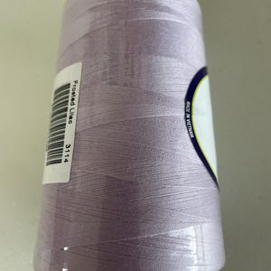 May include: A cylindrical spool of sewing thread in a light frosted lilac color. A white rectangular label is attached to the side, featuring a barcode, the text Frosted Lilac, and the number 3114. A partial label reads Made in Vietnam.