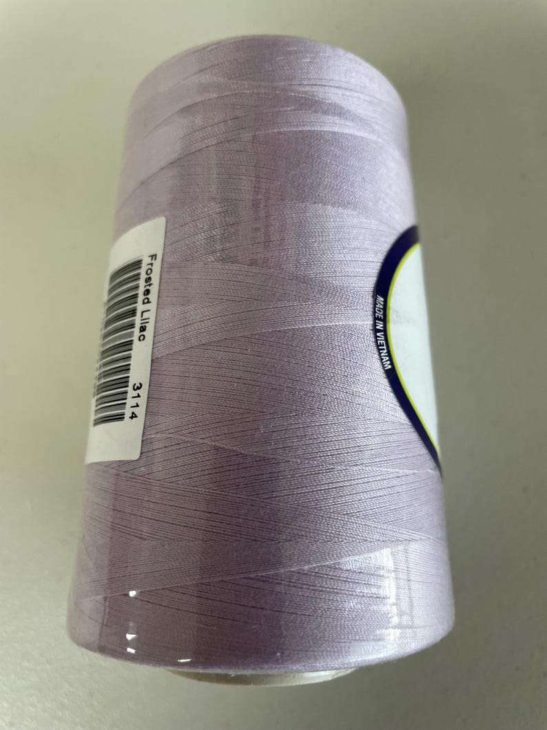 May include: A cylindrical spool of sewing thread in a light frosted lilac color. A white rectangular label is attached to the side, featuring a barcode, the text Frosted Lilac, and the number 3114. A partial label reads Made in Vietnam.