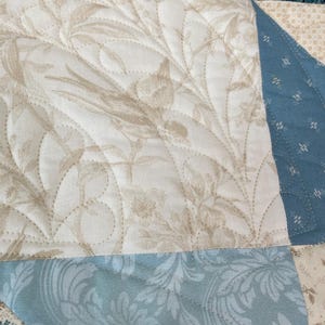 May include: Close-up of a quilted fabric with a floral pattern in beige and white. The quilt features a light blue section with a damask design and a dark blue section with a paisley pattern. The stitching is visible.
