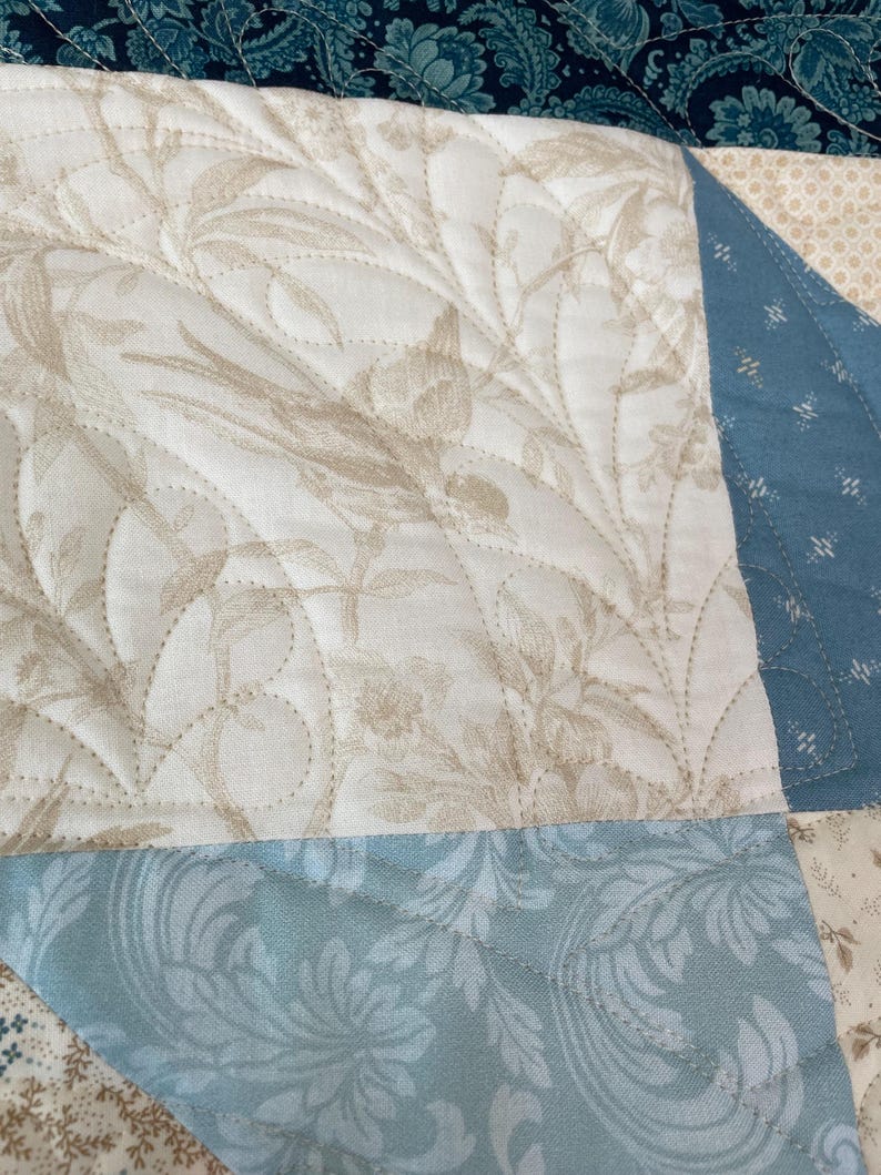 May include: Close-up of a quilted fabric with a floral pattern in beige and white. The quilt features a light blue section with a damask design and a dark blue section with a paisley pattern. The stitching is visible.