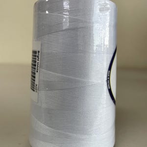May include: A large spool of bright white sewing thread wrapped in clear plastic film. A white label on the side features a barcode, the text Bright White, the number 3001, and the text Made in Vietnam printed along the curved edge of the label.
