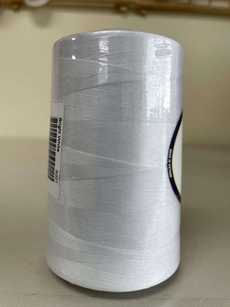 May include: A large spool of bright white sewing thread wrapped in clear plastic film. A white label on the side features a barcode, the text Bright White, the number 3001, and the text Made in Vietnam printed along the curved edge of the label.
