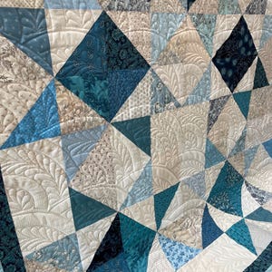 May include: A close-up of a handmade quilt with a geometric pattern. The quilt features various shades of blue, white, and cream fabrics. The quilt is machine quilted with intricate patterns, adding texture and detail to the design.