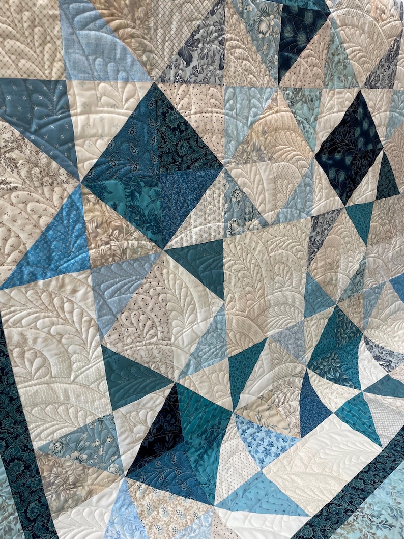 May include: A close-up of a handmade quilt with a geometric pattern. The quilt features various shades of blue, white, and cream fabrics. The quilt is machine quilted with intricate patterns, adding texture and detail to the design.