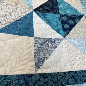 May include: Close-up of a handmade quilt with a patchwork design. The quilt features various shades of blue, white, and beige fabrics. The fabrics include floral and paisley patterns. The quilt is machine quilted with a decorative stitch.