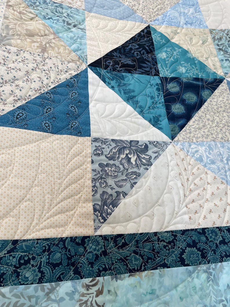 May include: Close-up of a handmade quilt with a patchwork design. The quilt features various shades of blue, white, and beige fabrics. The fabrics include floral and paisley patterns. The quilt is machine quilted with a decorative stitch.