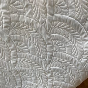 May include: Close-up of a white quilt with a repeating pattern of curved arcs and stylized feather designs. The quilt's texture is visible, with detailed stitching creating the raised patterns. The overall aesthetic is clean and elegant.