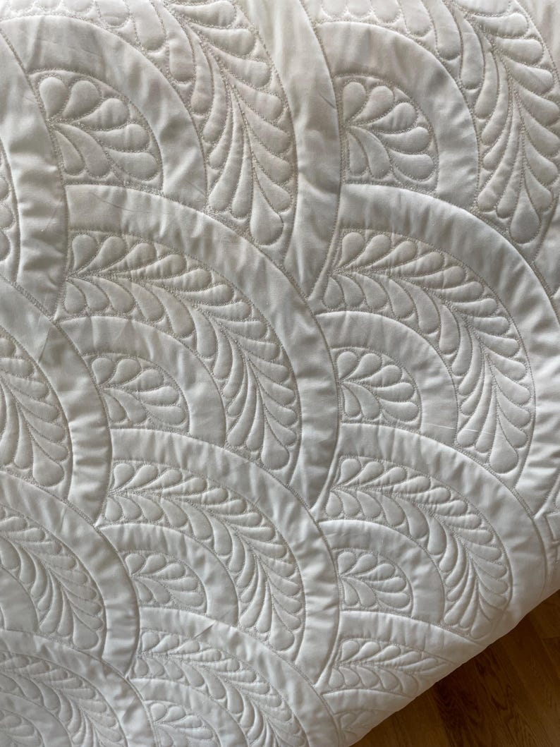 May include: Close-up of a white quilt with a repeating pattern of curved arcs and stylized feather designs. The quilt's texture is visible, with detailed stitching creating the raised patterns. The overall aesthetic is clean and elegant.
