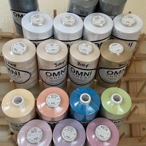 May include: A collection of fifteen cylindrical spools of Omni Thread arranged on a wooden rack. The spools feature various solid colors including white, gray, tan, peach, blue, light green, lavender, and pink, each with a label showing the color name.