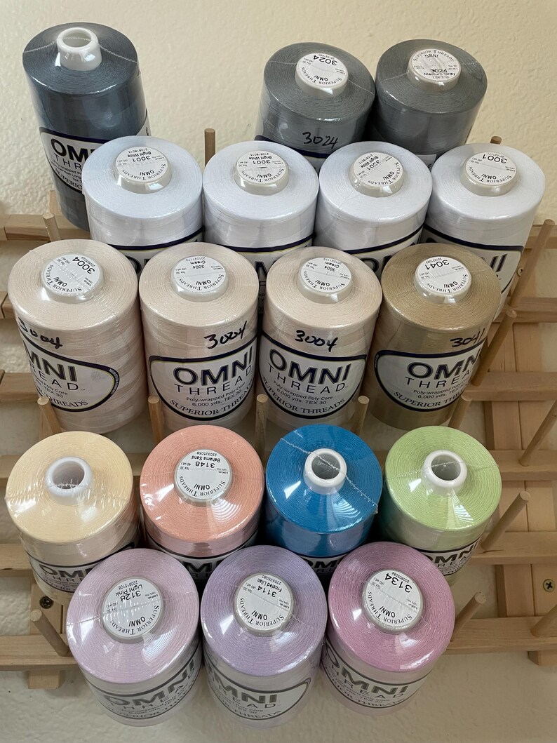 May include: A collection of fifteen cylindrical spools of Omni Thread arranged on a wooden rack. The spools feature various solid colors including white, gray, tan, peach, blue, light green, lavender, and pink, each with a label showing the color name.