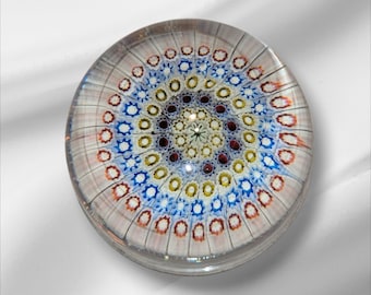 Puede incluir: Un pisapapeles redondo y transparente con un diseño intrincado y colorido. El patrón presenta círculos concéntricos de elementos azules, amarillos, rojos y blancos, creando una apariencia simétrica y floral. El pisapapeles es de vidrio.