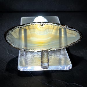 May include: A translucent, oval-shaped agate slice with cream, yellow, and white banded patterns. The stone features a dark, textured outer edge and is mounted on a clear acrylic stand with an integrated light source illuminating the center.