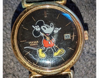 Könnte beinhalten: Eine goldfarbene Armbanduhr mit einem schwarzen Zifferblatt, auf dem ein Cartoon-Bild von Mickey Mouse abgebildet ist. Das Zifferblatt der Uhr enthält auch die Worte "Pulsar Quartz" und die Zahl "18".