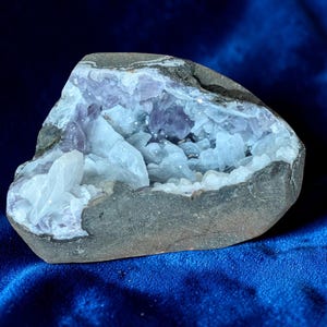 Può includere: Una geode con un esterno ruvido e grigio, che rivela una cavità piena di cristalli scintillanti, viola chiaro e bianchi. La geode è posta su uno sfondo blu scuro.