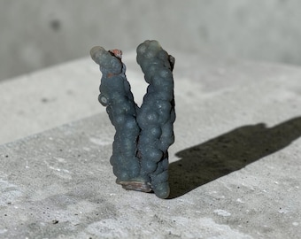 May include: A dark gray, botryoidal mineral specimen with a branched, vertical structure. The surface is composed of numerous small, rounded, bubble-like formations. The base is flat and brownish, while the upper sections split into two distinct, textured columns.