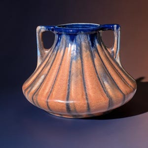 Könnte beinhalten: Eine Keramikvase mit breitem Boden und zwei Henkeln. Die Vase hat eine Farbgebung aus Orange und Blau mit vertikalen Streifen. Der obere Rand und die Henkel sind tiefblau, während der Körper orange mit dunkleren Streifen ist.