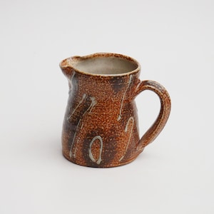 May include: A handmade ceramic jug with a speckled brown glaze and white drip accents. The jug has a rounded body, a curved handle, and a pouring spout. This rustic jug is ideal for serving beverages.
