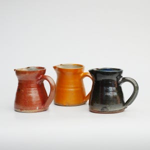 May include: Three handmade ceramic jugs in a row. The jugs are in shades of red, orange, and black. Each jug has a handle and a spout for pouring. The jugs are on a white background.