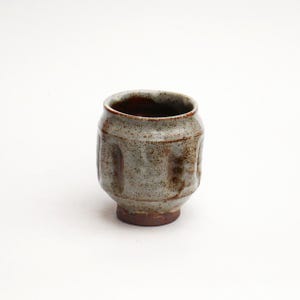 May include: A handmade ceramic cup with a speckled gray glaze and a dark brown interior. The cup has a rounded body with vertical indentations and a dark brown base. The cup is approximately 3 inches tall.