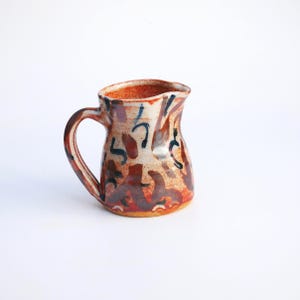Puede incluir: Una taza de cerámica blanca con un asa negra y el texto "But First, Coffee" en un estilo manuscrito. La taza está sobre una superficie blanca. La taza mide aproximadamente 10 cm de alto.