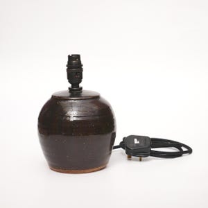 Könnte beinhalten: Eine dunkelbraune Keramik-Tischlampe mit schwarzem Lampenhalter. Die Lampe hat einen abgerundeten Körper und einen kleinen Sockel. Ein schwarzes Stromkabel und ein Stecker sind neben der Lampe zu sehen.