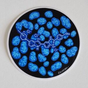 Op de afbeelding: Een ronde sticker met een zwarte achtergrond en een witte rand. De sticker toont een blauwe moleculaire structuur en blauwe celachtige vormen. De tekst "@SCIARTBRO" is rechtsonder zichtbaar.