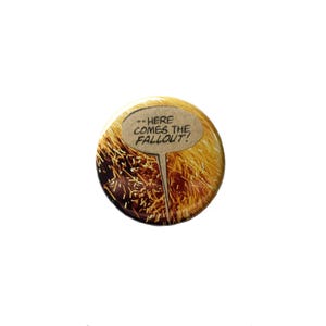 May include: A circular pin-back button featuring a graphic of golden sparks and dark shadows. A tan speech bubble is centered on the button with the black text "--HERE COMES THE FALLOUT!" printed inside. The design has a glossy finish.
