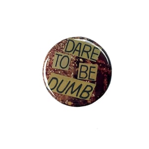 Puede incluir: Un botón redondo con un fondo marrón moteado y las palabras "DARE TO BE DUMB" en letras negras sobre rectángulos beige. El botón tiene un acabado brillante y está sobre un fondo blanco.