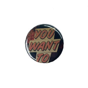 Puede incluir: Un botón redondo con las palabras "YOU WANT TO" en letras naranjas sobre un fondo beige. El botón tiene un borde negro y un acabado brillante. Es un botón de solapa.